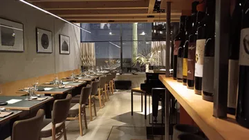 Modern restaurant interior with wooden tables and wine shelf.