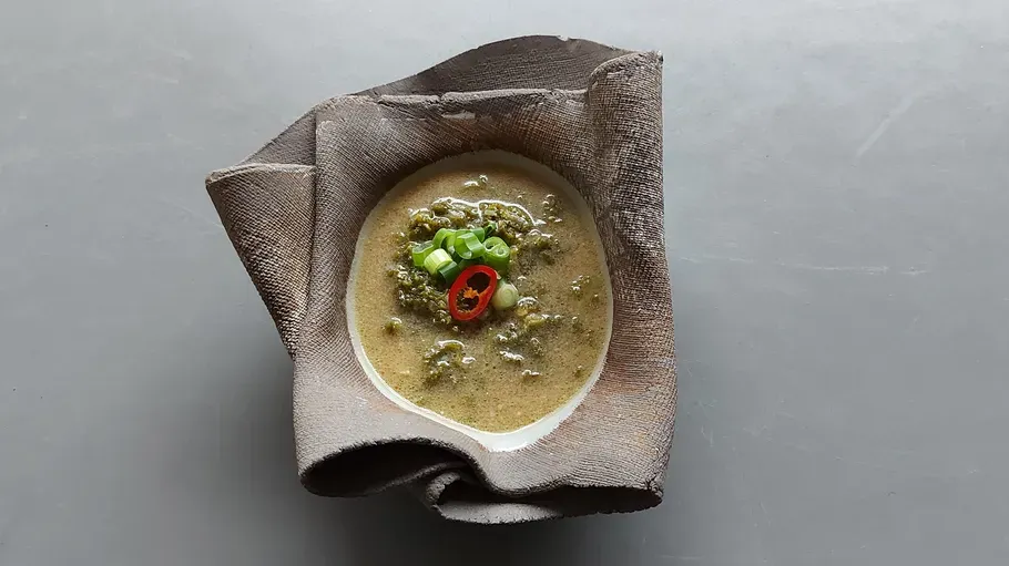 Schüssel mit grüner Suppe und Garnitur, grauer Hintergrund.