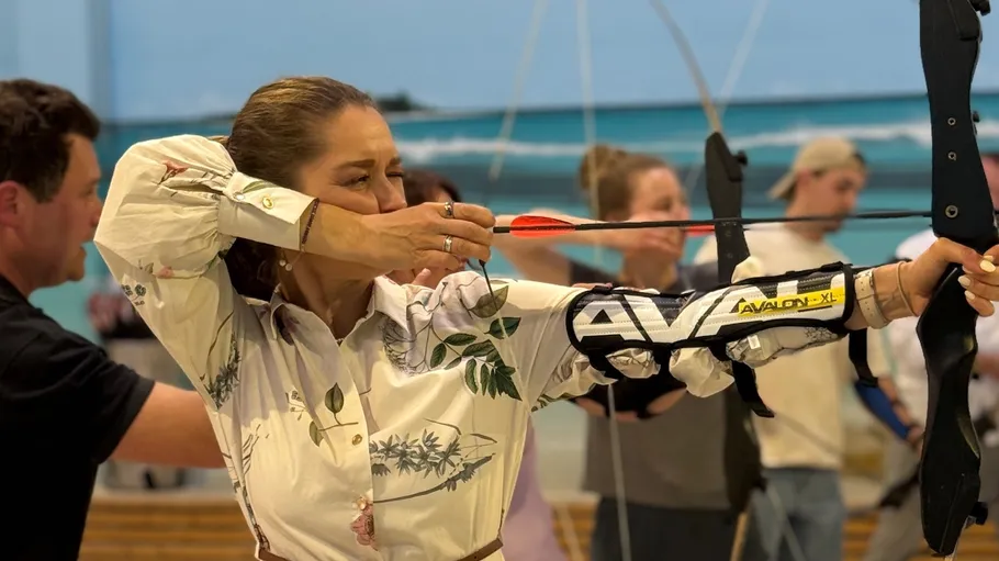 Woman aiming a bow indoors with people around.