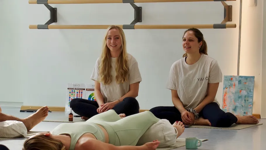 Zwei Frauen sitzen, eine weitere macht Yoga.