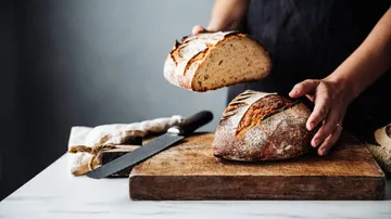 Person schneidet rustikales Brot auf Holzbrett.