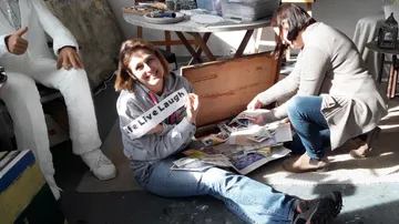 Frau lächelt, hält Schild, neben anderer ordnet Papiere.