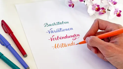 Hand schreibt bunte Wörter; Stifte und Orchideen sichtbar.