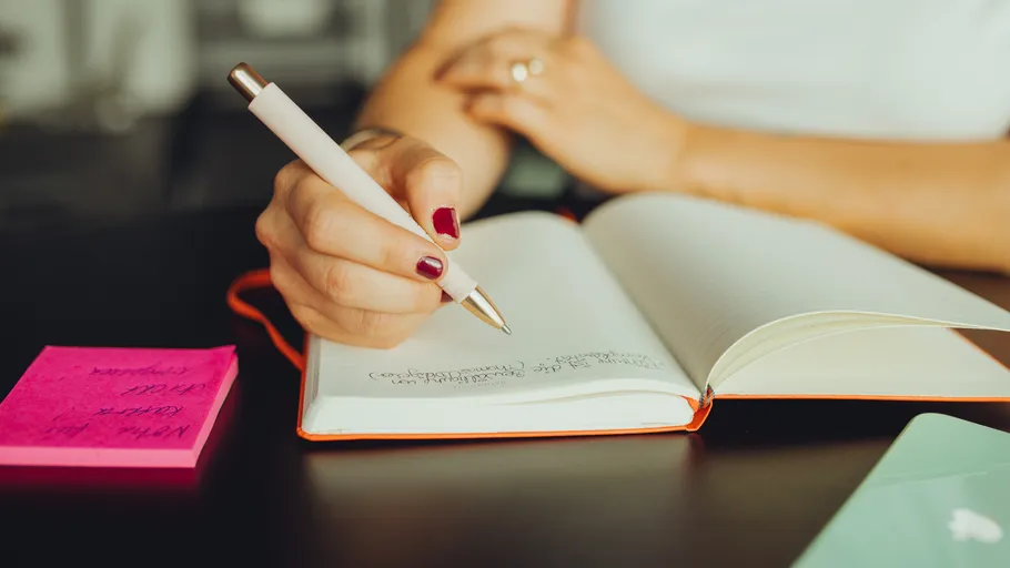 Person writing in notebook, pen poised, desk setting.