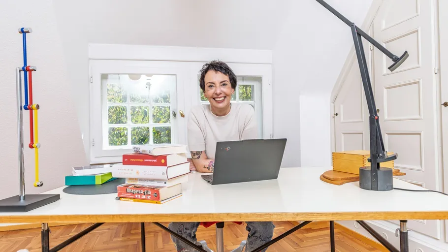Person smiling at desk with laptop, books, lamp.