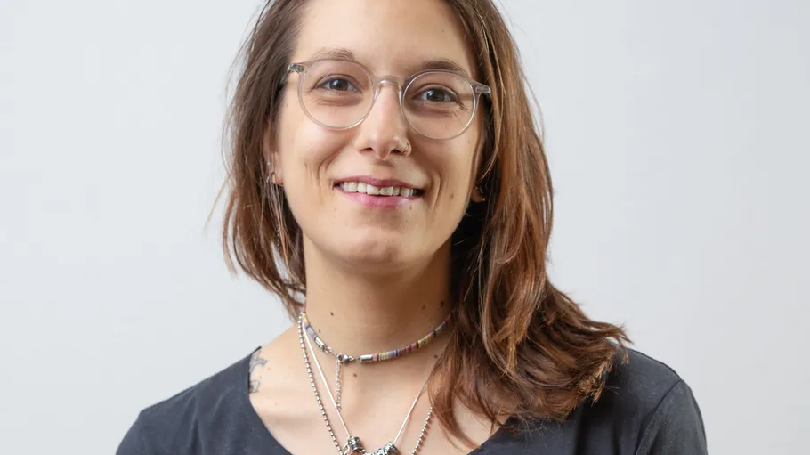 Smiling person wearing glasses and necklaces indoors.