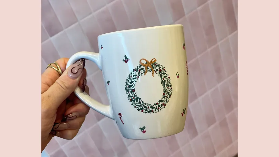 Decorated mug held by hand, pink background.