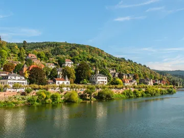 Fluss spiegelt Häuser am Ufer, umgeben von Hügeln.