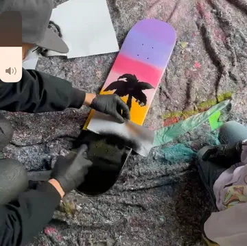 Person spray paints sunset skateboard design.