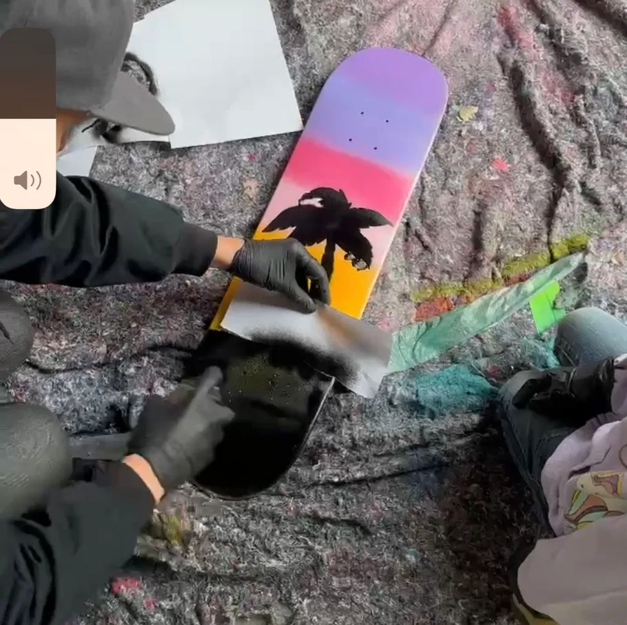 Person spray paints sunset skateboard design.