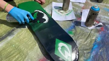 Person spray-painting skateboard with stencil design.