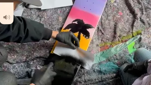 Person painting skateboard with stencil outside.