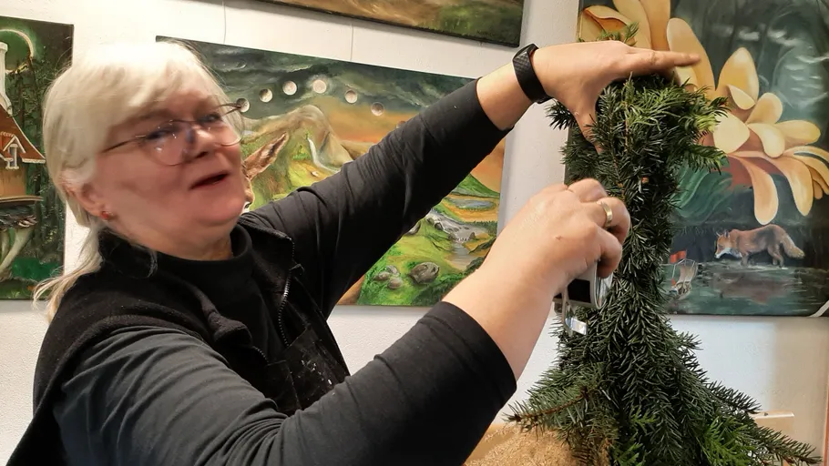 Woman shaping evergreen arrangement indoors, surrounded by paintings.