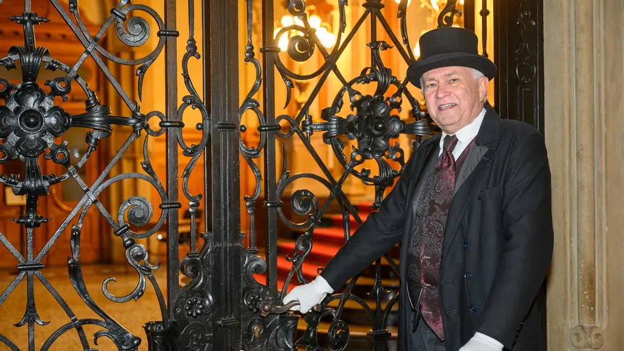 Man in top hat opens ornate gate indoors.