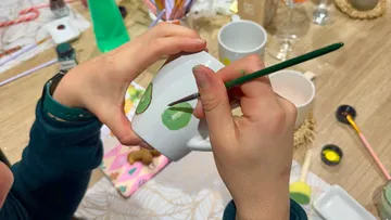 Hände malen grüne Muster auf weiße Tasse.