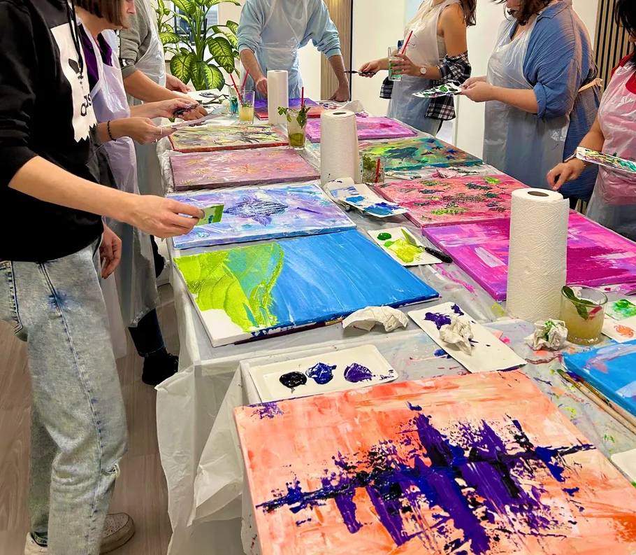 People painting colorful canvases on long table.