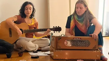Two women playing guitar and harmonium indoors.
