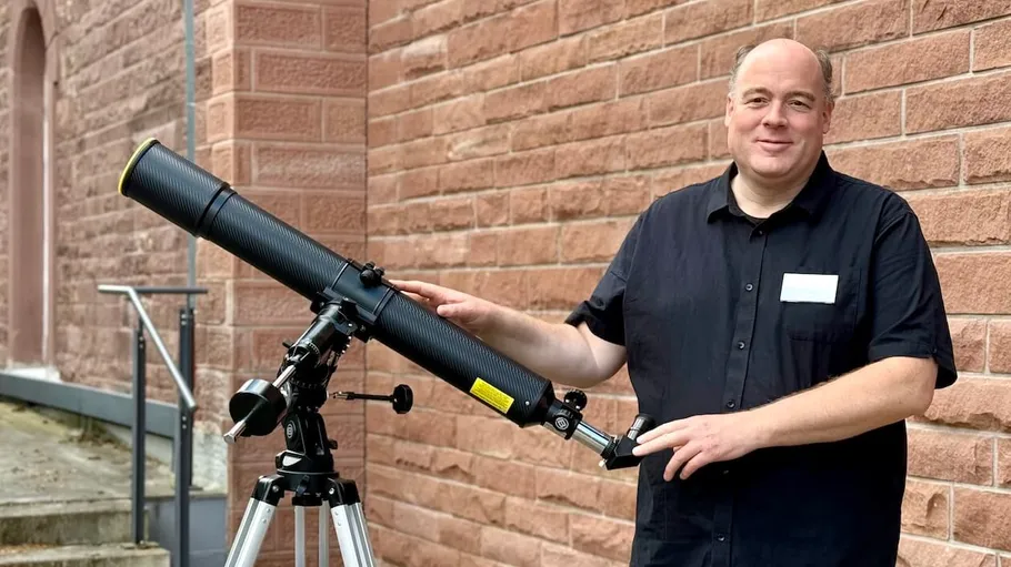 Man standing beside telescope near brick wall.