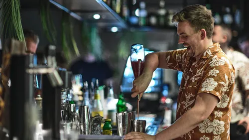 Barkeeper lächelt, macht Getränk an belebter Bar.