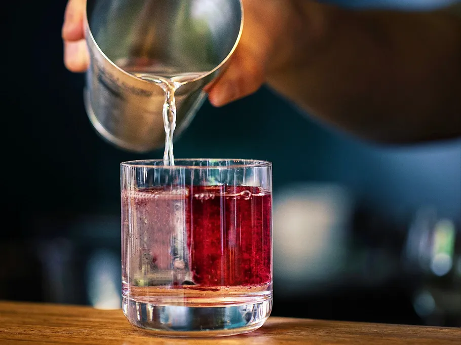 Hand pouring liquid into glass with cube.