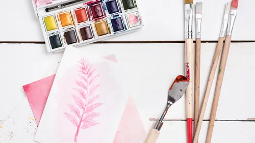 Watercolor paints, brushes, and pink painted paper.