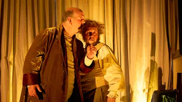 Two actors dressed in period costumes, dim lighting.