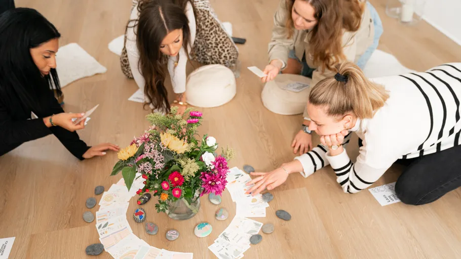 Four women sitting around floral arrangement, reaching for cards.