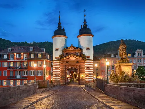 Tor mit Türmen erleuchtet, umgeben von Gebäuden und Bergen.