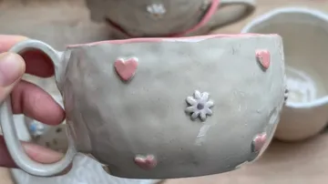 Hand hält Keramikbecher mit Herzdekorationen.