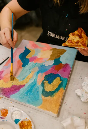 Person paints colorful canvas while holding pizza.