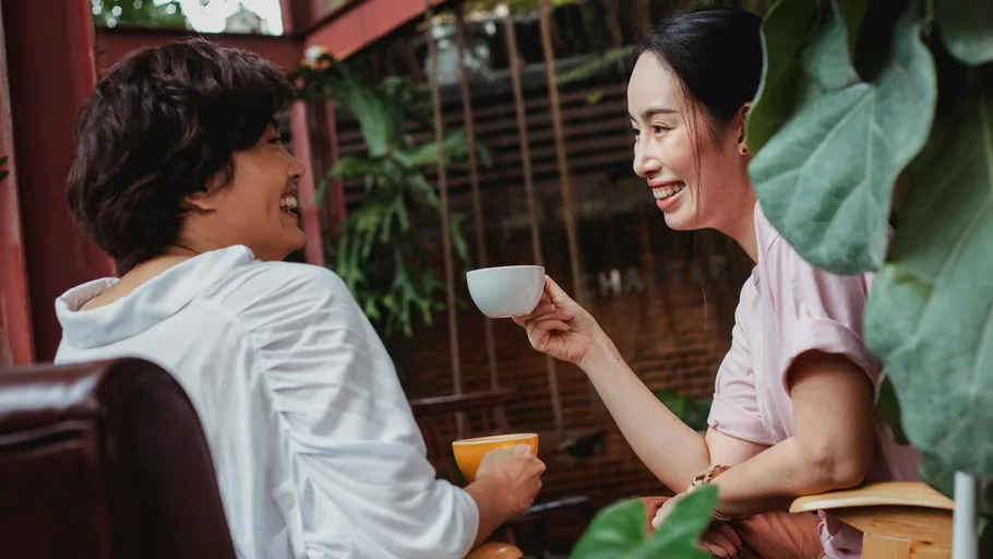 Two women smiling, holding cups, sitting outdoors.