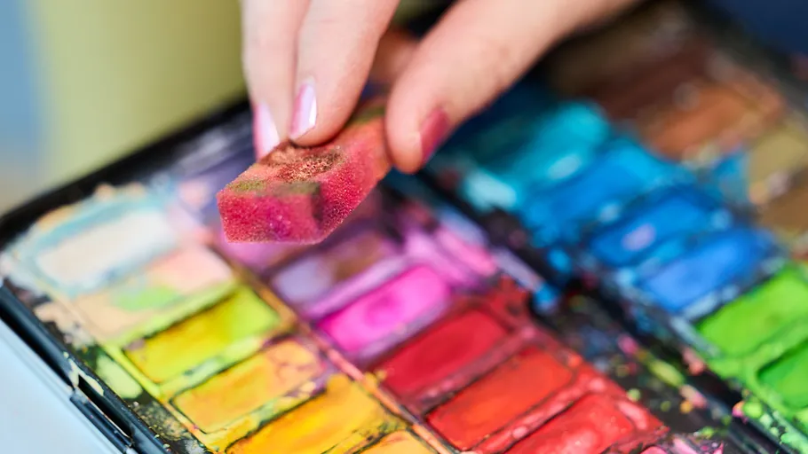Hand using sponge on colorful paint palette.