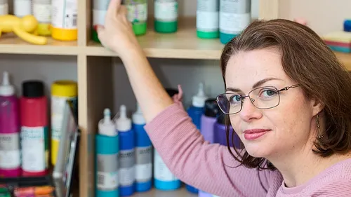 Woman reaching for paint bottle on shelf.