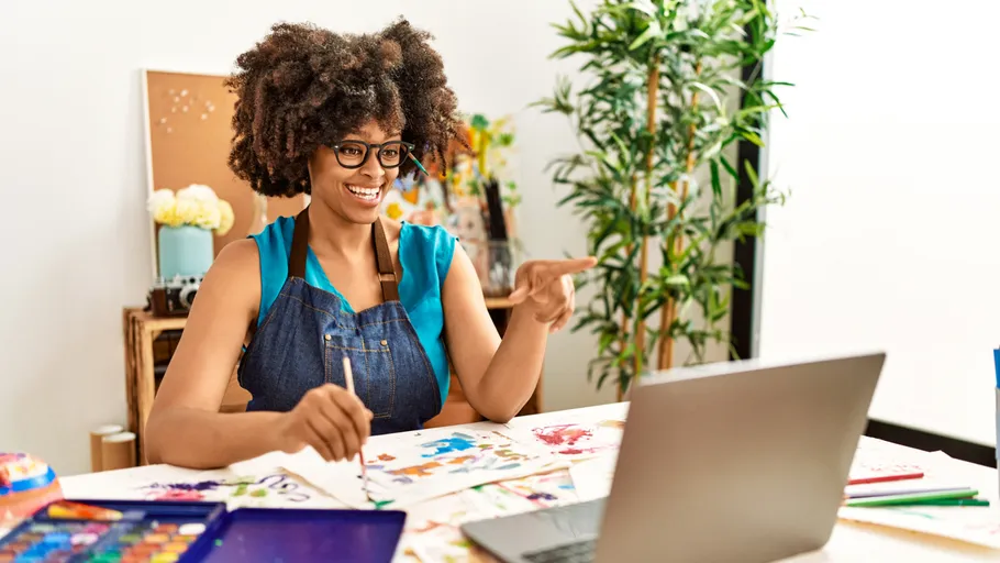 Woman painting at desk, smiling, using laptop.