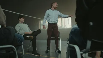 Man standing, speaking to seated group indoors.