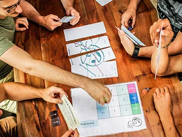 Several hands play at the table with cards and game materials.