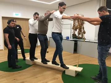Sechs Personen balancieren auf einem Holzbrett in einem Raum.