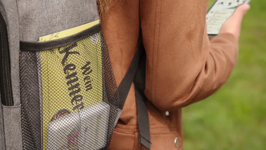 Rucksack mit Buch, Person hält Handy, draußen.
