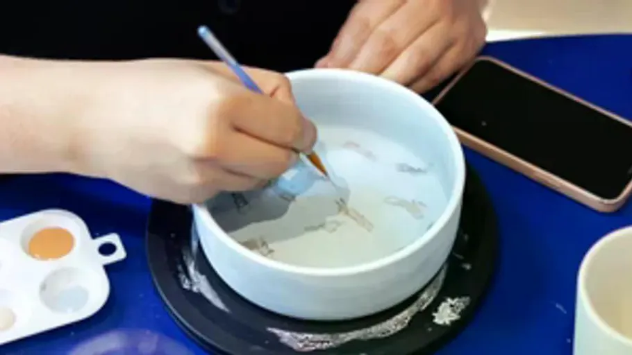 Person painting bowl at table with phone.