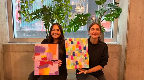 Two women sitting, displaying colorful paintings.