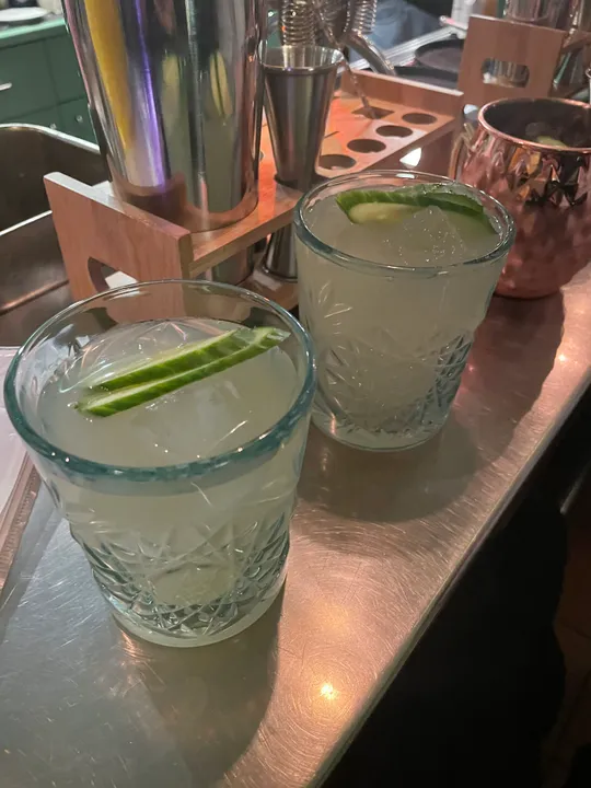 Two cocktails with cucumber on a bar.