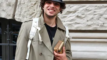 Mann hält Bücher, lächelt, trägt Trenchcoat.