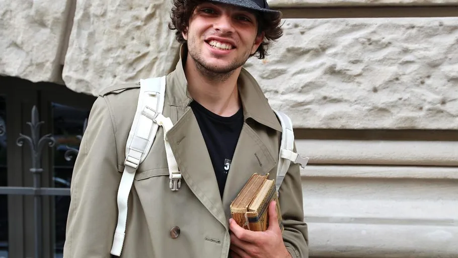 Mann hält Bücher, lächelt, trägt Trenchcoat.