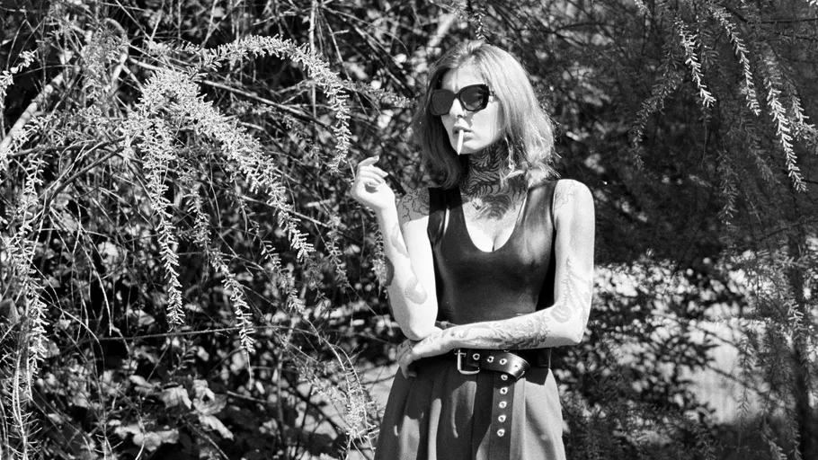 Woman with tattoos smoking, standing among bushes.
