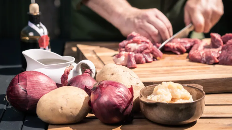 Person schneidet Fleisch neben Zwiebeln und Kartoffeln.