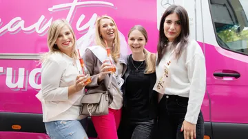 Vier Frauen lächeln mit Getränken vor pinkem Van.