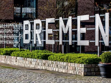 Großes `BREMEN`-Schild steht vor einer Backsteinmauer, umlagert von Pflanzen.