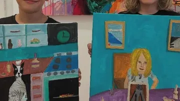 Two children holding colorful paintings indoors.