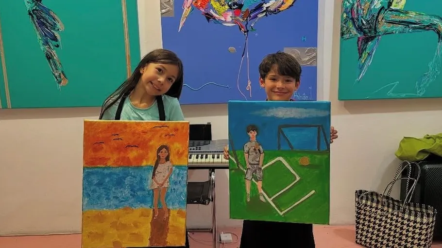 Two children proudly hold colorful paintings indoors.