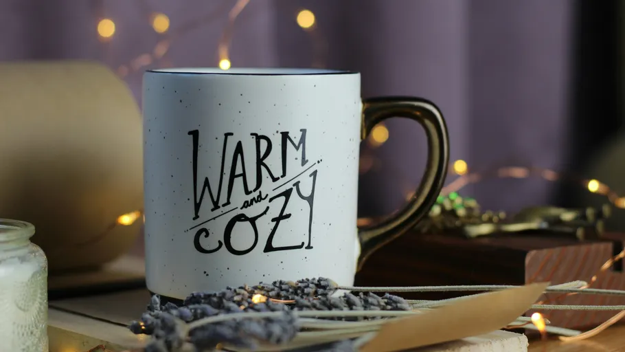 Tasse mit 'Warm and Cozy' Text, Lichter im Hintergrund.